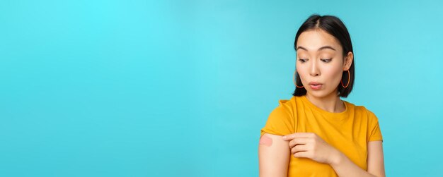 Vaccine campaign from covid19 Young beautiful healthy asian woman showing shoulder with bandaid concept of vaccination standing over blue background