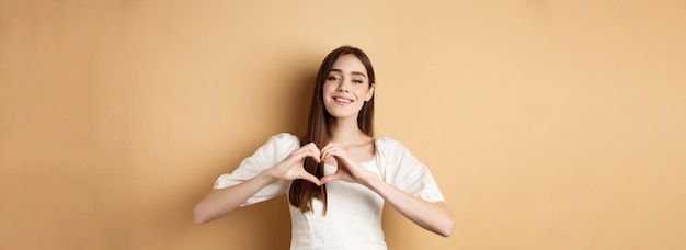 Free photo valentines day attractive young girl in white dress smiling and showing heart gesture standing on be
