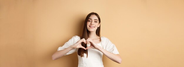 Free Photo valentines day attractive young girl in white dress smiling and showing heart gesture standing on be