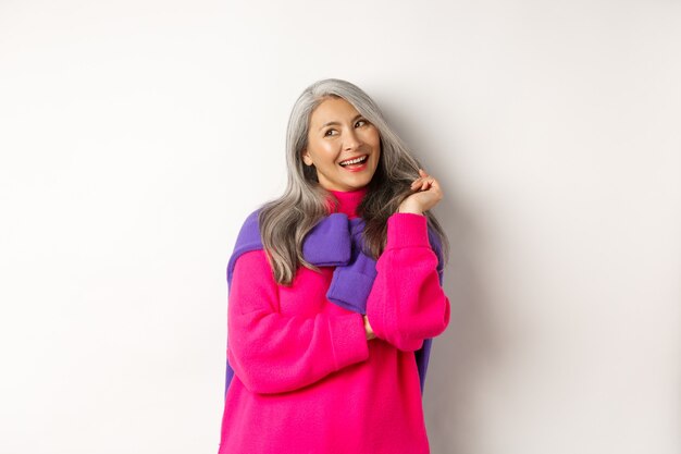 Valentines day concept. Beautiful asian senior woman laughing coquettish, playing with hair strands and looking at upper left corner flirty, standing over white background