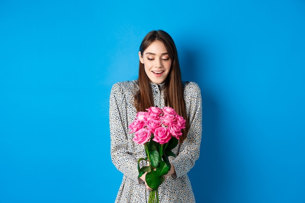 バレンタインデーのコンセプト。幸せな魅力的な女性は、青い背景の上に立って、ピンクのバラの花束に感謝している驚きの花を受け取ります。