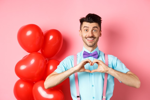 Free photo valentines day concept romantic guy in bowtie showing heart gesture and say i love you with hopeful ...