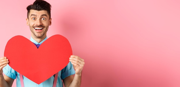 Foto gratuita concetto di giorno di san valentino uomo romantico che si innamora mostrando un grande ritaglio di cuore rosso e standin sorridente