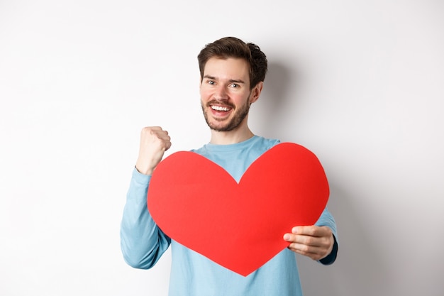 Valentines day. Happy boyfriend triumphing, saying yes and showing valentine red heart, smiling as winning girls love, standing over white background