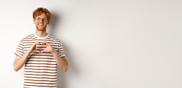 Free photo valentines day happy boyfriend with red hair smiling and showing heart gesture i love you standing o