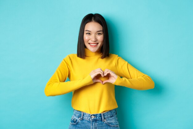 Valentines day and romance concept. Beautiful asian woman show I love you, heart gesture and smiling, standing against blue background