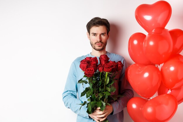 Valentines day romance. Confident young man holding bouquet of red roses, standing near hearts balloons, going on romantic date with lover, white background