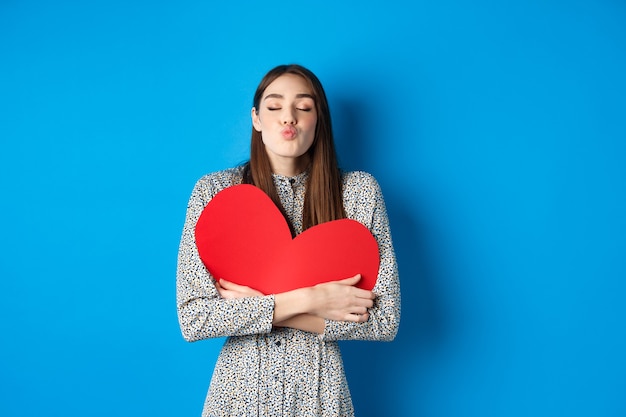 Valentines day. Romantic beautiful woman close eyes and pucker lips for kiss, holding big red heart cutout, kissing you, standing on blue background.