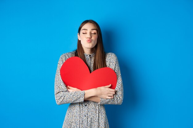 Valentines day. Romantic beautiful woman close eyes and pucker lips for kiss, holding big red heart cutout, kissing you, standing on blue background.