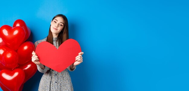 Valentines day romantic girl in dress showing big red heart cutout dreaming of love standing near ho