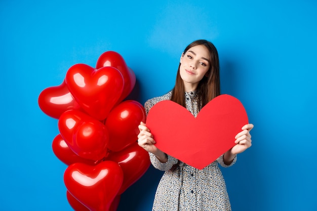 Valentines day. Romantic girl in dress showing big red heart cutout, dreaming of love, standing near holiday balloons on blue background