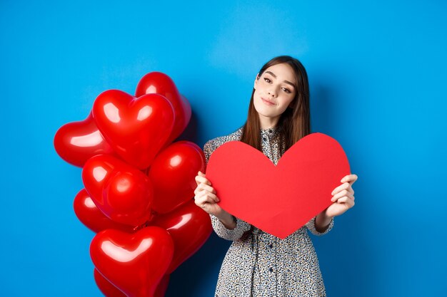 Valentines day. Romantic girl in dress showing big red heart cutout, dreaming of love, standing near holiday balloons on blue background