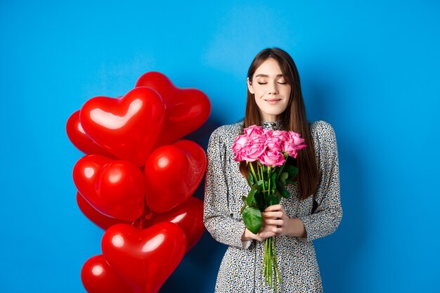 Valentines day. Romantic pretty woman close eyes and smelling beautiful flowers, standing near heart balloons, blue background