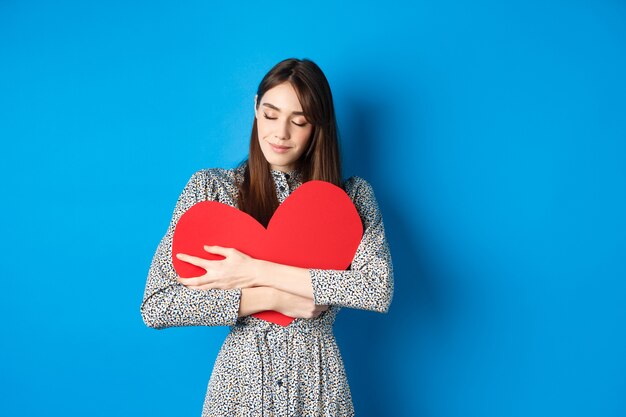 Valentines day romantinc girl in dress hugging big red heart cutout close eyes and smile with dreamy...
