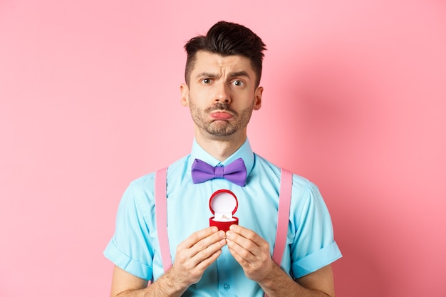 Valentines day. Sad boyfriend being rejected, showing engagement ring and sulking upset, she said no, standing over pink background.