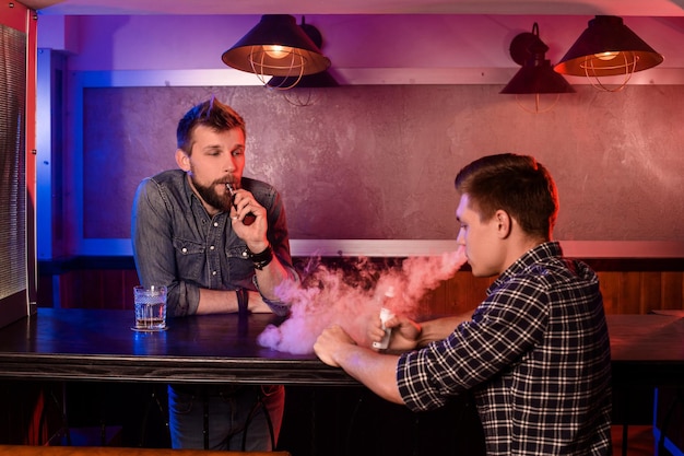 Вейпинг мужчина держит мод. Облако пара в вейпбаре. Двое мужчин отдыхают в баре и курят электронные сигареты.