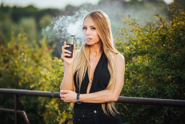 Vaping. Young beautiful woman smoking e-cigarette with smoke outdoors. 