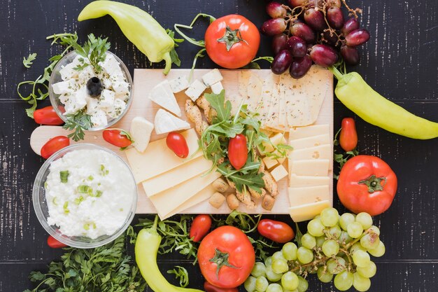 Variety of cheese slices and cubes with grapes, tomatoes; green chilies; arugula leaves and parsley on black background