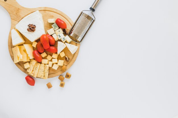 Variety of cheese with red tomatoes on chopping board with grater on white backdrop