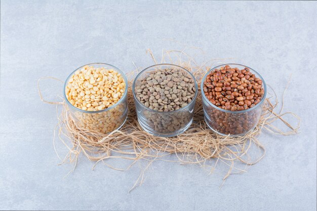 Variety of legumes assorted in glass bowls on a pile of straw on marble background.