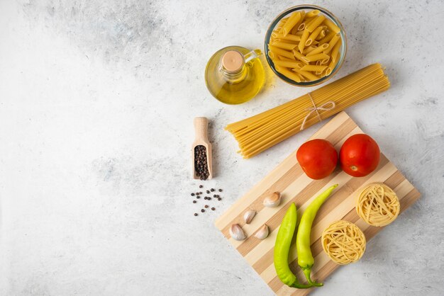 Variety of raw pasta, bottle of olive oil, pepper grains and vegetables on white table. 