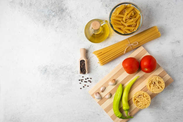 Variety of raw pasta, bottle of olive oil, pepper grains and vegetables on white table. 