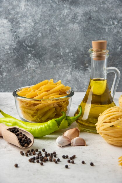 Разнообразие сырых макарон, бутылки оливкового масла, зерен перца и овощей на белом столе.