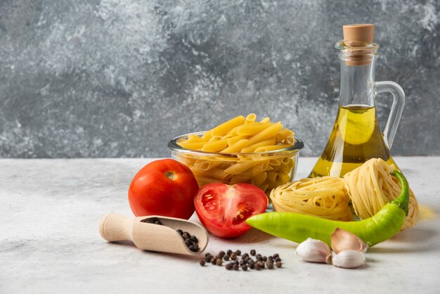 Variety of raw pasta, bottle of olive oil, pepper grains and vegetables on white table. 