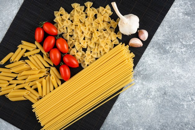 Variety of raw pasta and vegetables on black bamboo sheet.