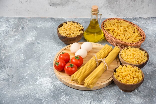 Variety of raw pasta with eggs, fresh tomatoes and bottle of oil on marble surface.