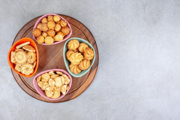 Various bowls of crispy biscuits and cookie chips on wooden board on marble surface