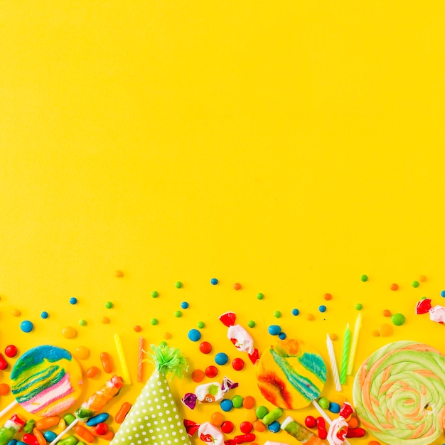 Various candies and party hat at the bottom of yellow background