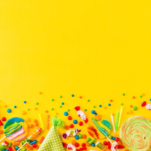 Various candies and party hat at the bottom of yellow background