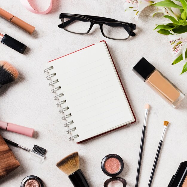 Various cosmetics and glasses scattered around blank notebook
