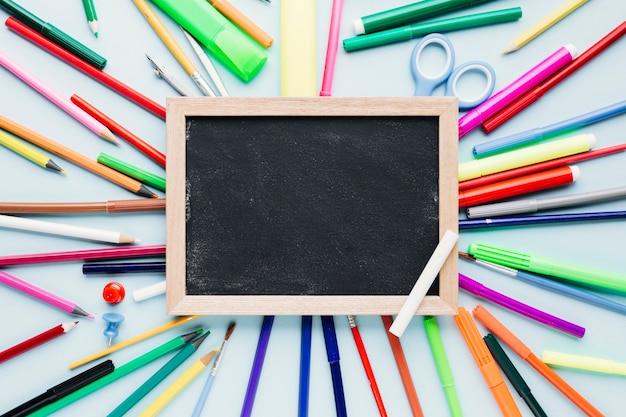 Various drawing tools scattered around blank chalkboard on blue desk