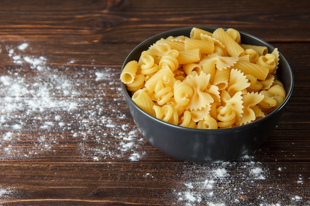 Various dry pasta with sprinkled flour in a bowl on wooden table, high angle view.