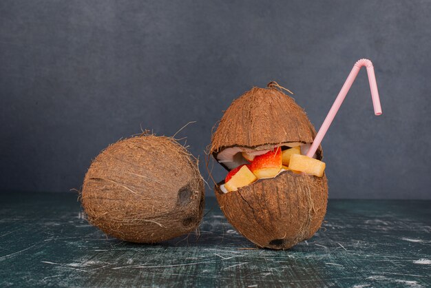 Various fruits in half cut coconut with straw on marble table.