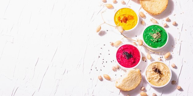 Various hummus dips, the flat lay of hummus in different colors with spinach, beetroot, turmeric and vegetables, vegan snack. white putty background, top view, banner