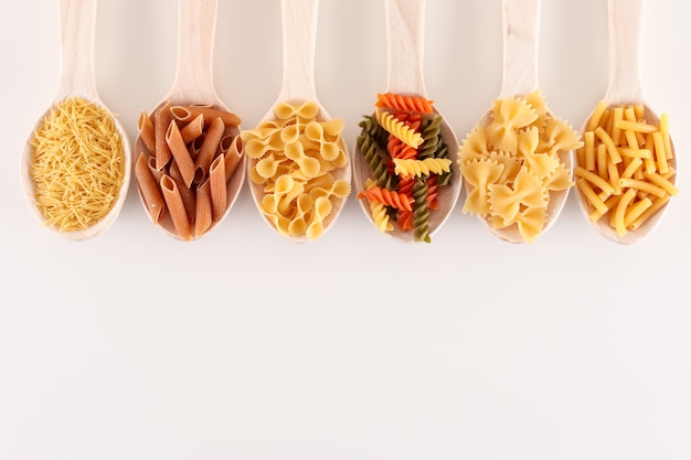 various kind of pasta in wooden spoons on white surface with copy space