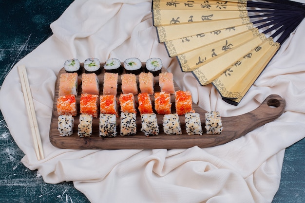 Различные виды суши-роллов подаются на деревянном блюде с палочками для еды и японским веером.