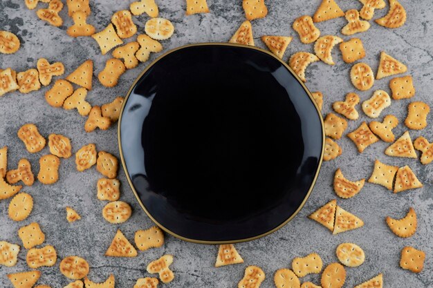 Various small crackers scattered near black plate on marble background.