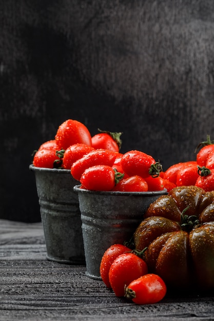 Various tomatoes in mini buckets on grey wooden and dark wall, side view.