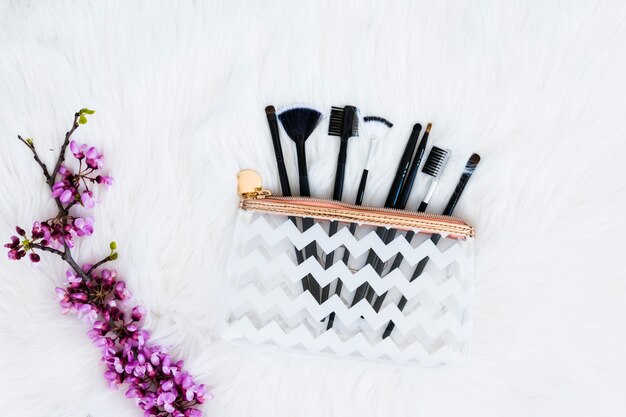 Various type of makeup brushes in plastic transparent bag with purple flower twig on white fur