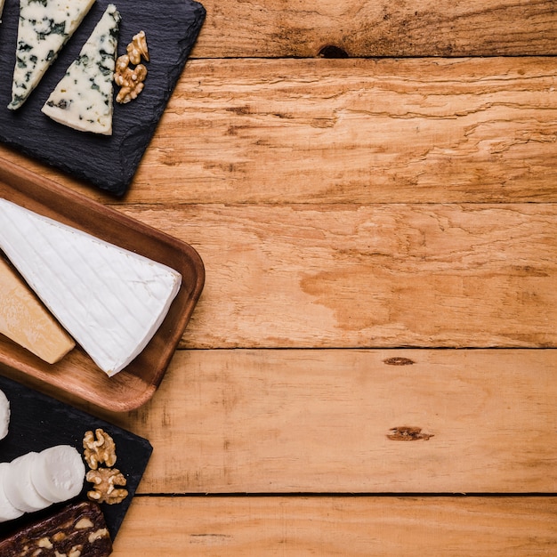 Various types of fresh cheese pieces on wooden plate and stone tray over wooden backdrop