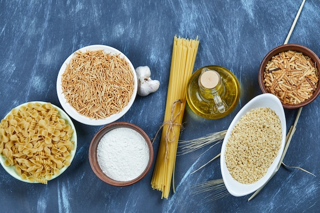 Various uncooked pasta with a bottle of oil, flour and garlic.