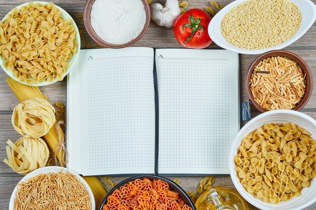 Various uncooked pasta with notebook and vegetables on a wooden table.