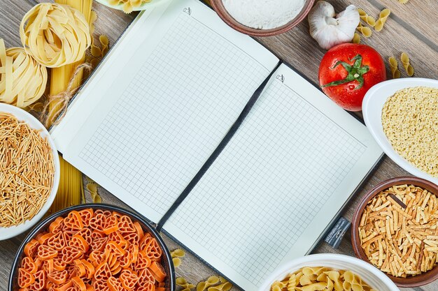 Various uncooked pasta with notebook and vegetables on a wooden table.