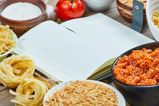 Various uncooked pasta with notebook and vegetables on a wooden table.