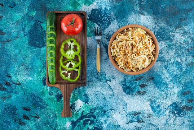 Variously vegetables on a board next to fork an a bowl of noodle on the blue table. 