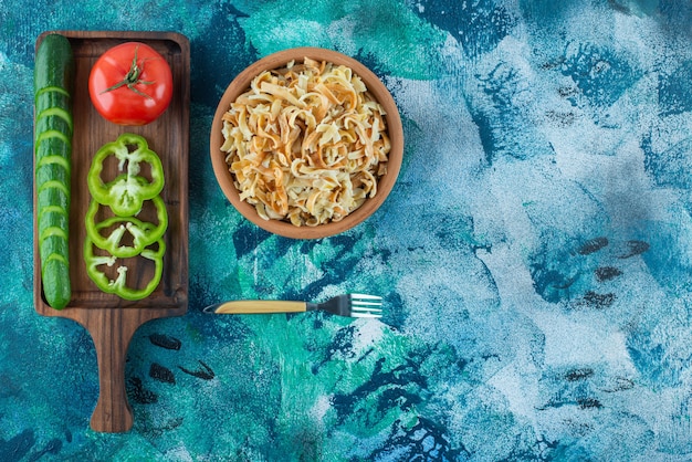 Variously vegetables on a board next to fork an a bowl of noodle on the blue table. 